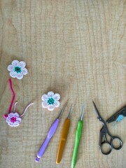 Crochet needles, scissors and crochet flowers on a light brown wooden background.