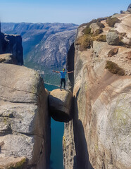 A young man wearing blue T-shirt standing on famous rock, Kjerag, Norway. The ball-shaped rock is...