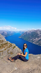 Naklejka na ściany i meble A girl wearing blue T-shirt sitting at the edge of a steep cliff of Preikestolen, with a view on Lysefjorden. Fjord goes far inland. Girl enjoys the view, feeling free and happy. Great accomplishment