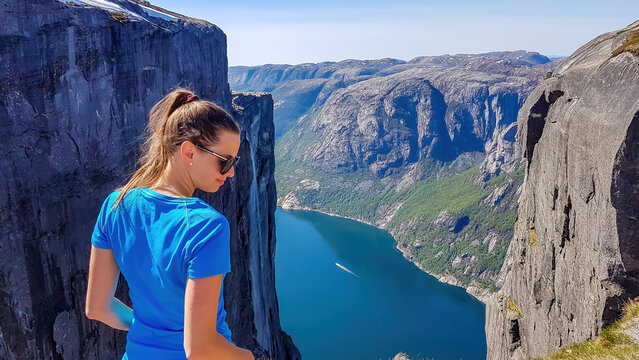 Girl wearing blue T-shirt looks down, past her shoulder. She is enjoying the fjord view in front of her. Stunning Lysefjorden shimmers with many shades of blue and green. Girl is happy after the hike