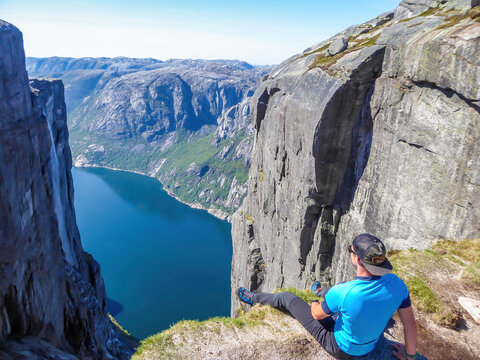 A young man sitting at the edge of a steep mountain. His legs are hanging down. Overcoming the fear of heights. In front of her stunning Lysefjorden shimmering with many shades of blue and green