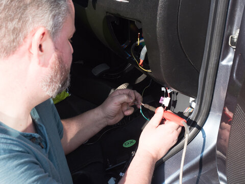 A bearded male mechanic works on the wiring of a motorhome recreational vehicle in front footwell with a soldering iron.