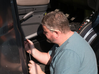 A male bearded mechanic works in the front cab footwell of a motorhome recreational vehicle.He has a pencil behind an ear and screwdriver in hand