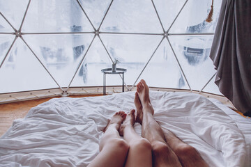 Legs of a man and woman couple in bed against the background of a snow-covered forest in a dome camping. © Kathleen