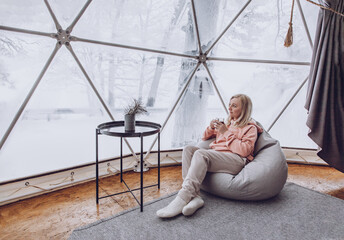 A woman is sitting in a geo dome glamping tent in a bag chair and looking out at the winter...