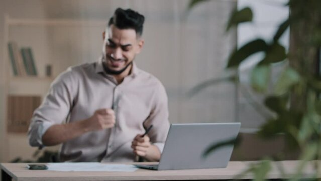 Blurred view with camera autofocus hispanic man joyful businessman dancing at workplace celebrating good news pretend playing drums having fun enjoying work break listen music singing song at office
