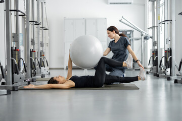 Woman doing exercises with fitness ball with rehabilitation specialist at the gym. Concept of physical therapy for health and recovery. Idea of recovery after pregnancy - obrazy, fototapety, plakaty