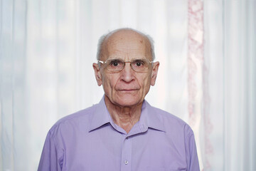 Portrait of hoary elderly man wearing eyeglasses standing at home against white transparent curtain