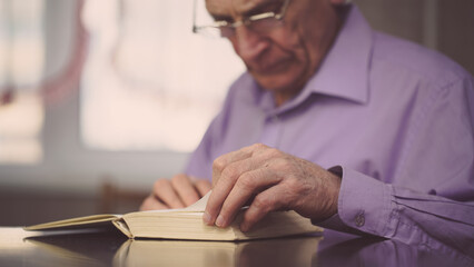 Clever mature professor in eyewear seated at wooden table and reads book at home