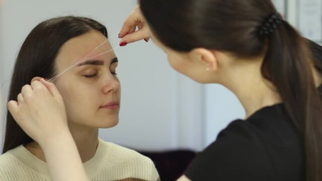 professional correction eyebrows shape, removing unwanted hair using thread, threading brows beauty salon. beautician stylist makes eyebrows removing with string. caucasian female close-up face brows