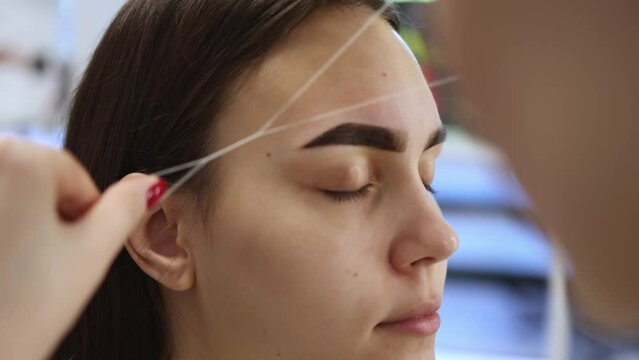 professional correction eyebrows shape, removing unwanted hair using thread, threading brows beauty salon. beautician stylist makes eyebrows removing with string. caucasian female close-up face brows