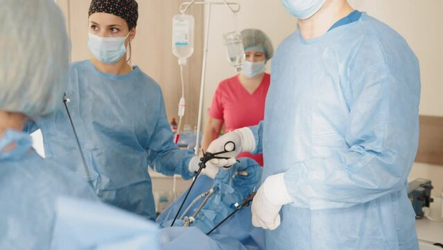Portrait of female medical worker in protective mask and sterile gloves holding laparoscopic grasper with trocar while standing near table with surgical tools. Surgeon and his assistant on blurred.