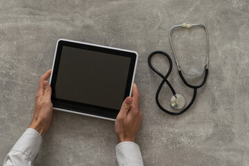 Doctor hand holding tablet with blank screen and stethoscope on grey table, copy space, top view