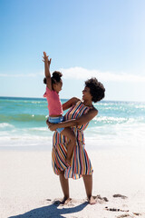 Cheerful african american mother carrying daughter while enjoying sunny day at beach against sky