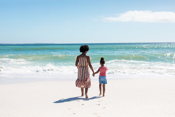 Rear view of african american mother and daughter holding hands while walking towards sea at beach