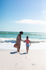 African american mother holding hand of daughter while walking towards sea at beach on sunny day