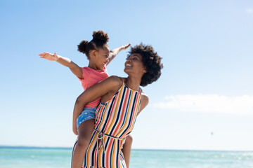 Happy african american woman looking at daughter with arms outstretched on her back at beach