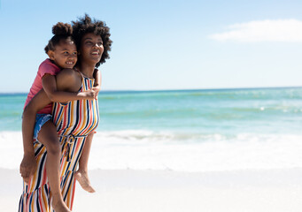 Smiling african american mother giving piggyback ride to daughter at beach on sunny day
