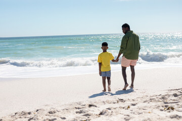 Full length rear view of african american father and son standing at beach on sunny day
