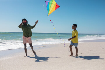 Full length of african american father and son running with kite at beach against clear blue sky