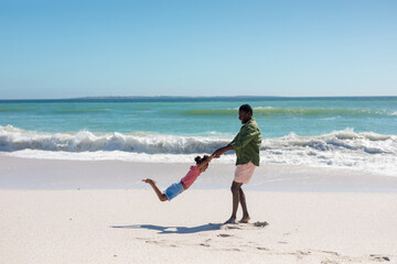 Happy african american man spinning daughter at beach while enjoying summer holiday together