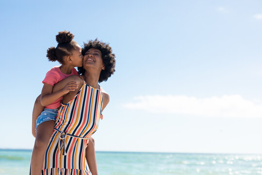 African american girl kissing mother while enjoying piggyback ride on her at beach against blue sky