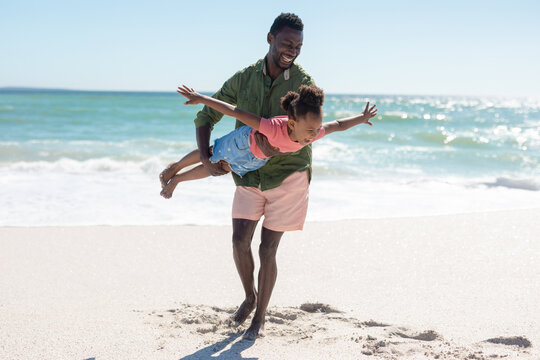 Full Length Of Happy African American Man Carrying Daughter Pretending To Fly At Beach On Sunny Day