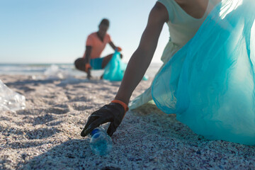 Midsection of african american woman picking plastic bottle from sand at beach