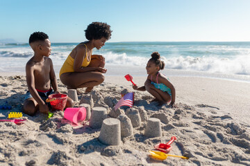 Fototapeta premium African american mother crouching by children making sandcastles at beach on sunny day