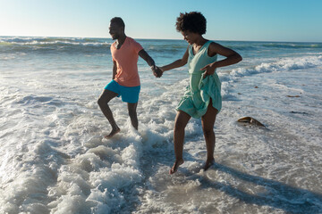 Happy african american couple enjoying summer holiday wading on shore at beach during sunny day