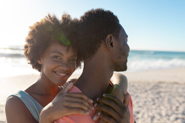 Portrait of smiling african american woman hugging boyfriend from behind at beach on sunny day