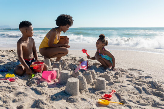 African american mother crouching by children making sandcastles at beach on sunny day