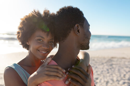 Portrait Of Smiling African American Woman Hugging Boyfriend From Behind At Beach On Sunny Day