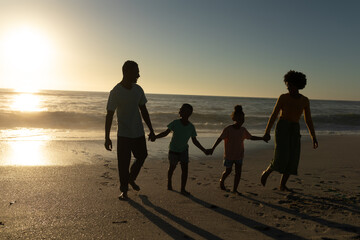Full length of african american family holding hands walking at beach against sky during sunset - Powered by Adobe