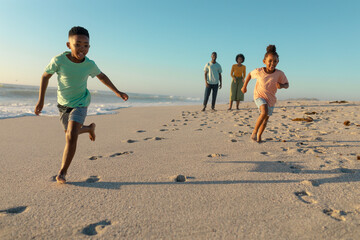 Full length of happy african american children running against parents walking at beach on sunny day