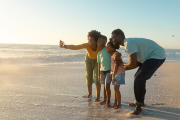 Happy african american mother taking selfie with family on shore at beach during sunset