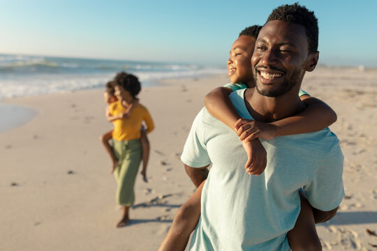 Happy african american man piggybacking son with mother and daughter walking in background at beach