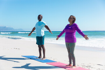Full length of african american retired senior couple practicing yoga at beach against blue sky