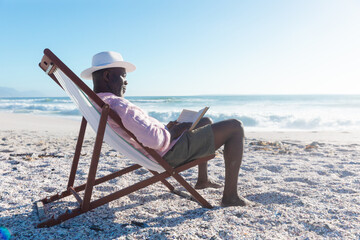 Side view of african american senior man reading book sitting on folding chair at beach