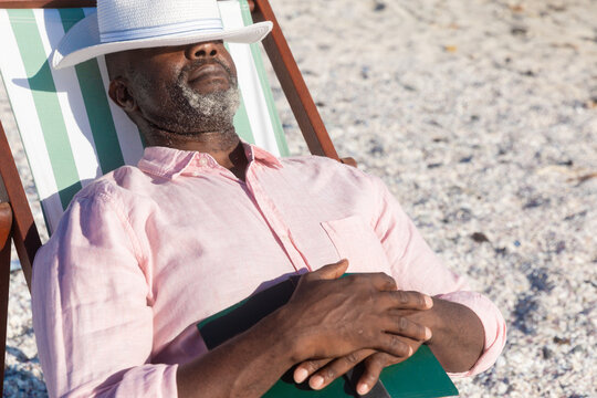 Retired african american senior man sleeping with book and hat over eyes on folding chair at beach