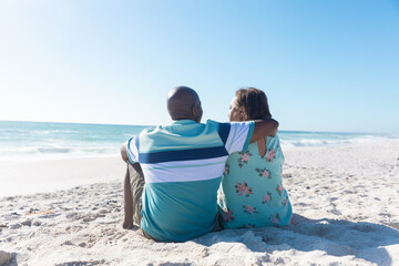 Retired african american senior couple sitting on sand at beach with copy space over blue sky