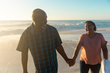 Happy african american senior couple holding hands walking together at beach during sunset - Powered by Adobe