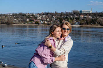 Portrait of smiling happy senior mother with her adult daughter hugging, having fun and walking on a sandy beach by the river.