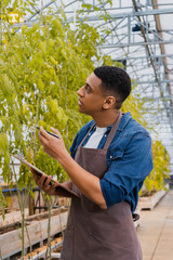 Side view of african american farmer holding pen and clipboard near plants in greenhouse.
