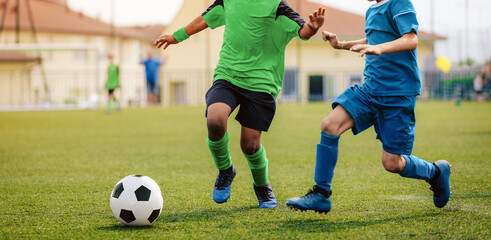 Obraz premium Two soccer players kicking soccer ball in a game. School boys play sports competition. Two multiracial kids playing football match. Kids in green and blue soccer jersy uniforms