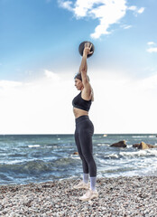 Fototapeta na wymiar Strong athletic woman exercising with medicine ball on the beach during the day with blue sky and clouds. Functional outdoor training