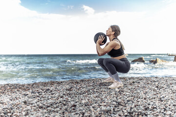 Strong athletic woman exercising with medicine ball on the beach during the day with blue sky and clouds. Functional outdoor training
