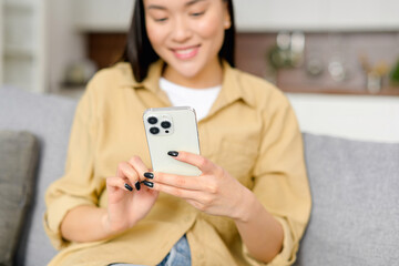 Selective focus at smart phone in female hands. Cropped view of the asian woman sitting on couch using smartphone, spending time online, texting with friends, scrolling news feed in social networks