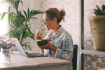 Side view of adult young woman smiling and eating in front of a laptop at the desk at home....