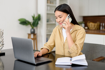 Girl works remotely. Multitask asian freelance woman talks on the smartphone and takes notes with pen, using laptop for remote work from home, write down some information sitting at the table